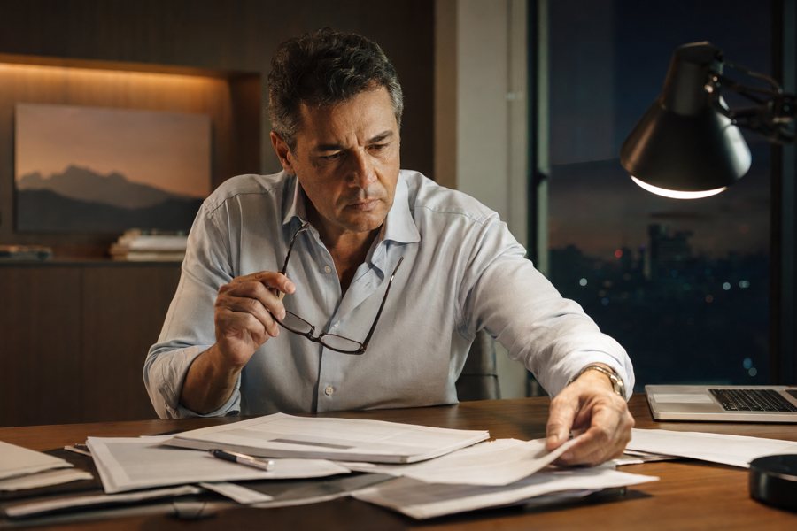 Empresário preocupado revisando contratos em papel à noite, sem conseguir localizar uma prova